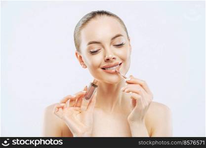 Photo of tender woman with perfect makeup, applies lip gloss, uses lip concealer brush, has dark combed hair, stands bare shoulders indoor, cares about herself, isolated on white background.