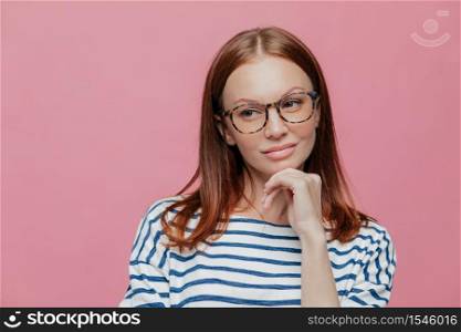 Photo of thoughtful attractive young woman holds chin, wears spectacles, casual clothes, contemplates about something, models over pink background with free space for your text or advertisement