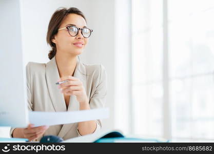 Photo of thoughtful brunette woman in formal wear, spectacles, holds pen and paper, looks pensively away, thinks about development of own business, works freelance, poses in coworking space.