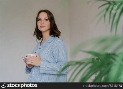 Photo of thoughtful young woman with dark hair, appealing appearance, warms with hot beverage, dressed in domestic costume, poses indoor near green plant, blank space for your advertisement.