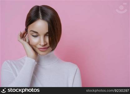 Photo of thouhtful shy young woman looks down, has dreamy expression, dressed in casual clothing, thinks about something pleasant, isolated over rosy background, blank space for your promotion