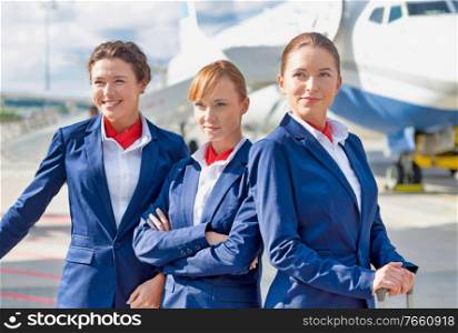 Photo of three confident flight attendants standing against airplane in airport