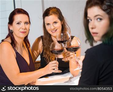Photo of three female friends toasting with red wine in the kitchen. Focus on middle woman.&#xA;