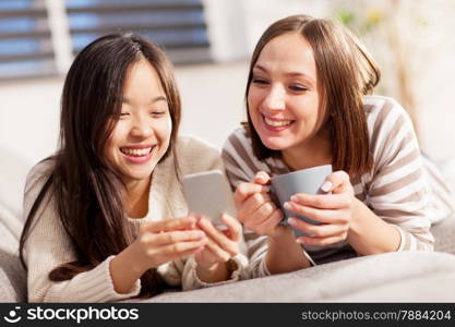 Photo of two smiling girls lying on the sofa in living room and reading a text message on the phone