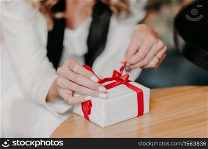 Photo of unrecognizable Caucasian woman ties red ribbon on small white gift box, prepares surprise for somebody on Chirstmas, New Year or birthday, blurred background. Close up, selective focus