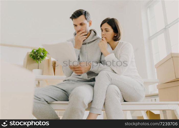 Photo of upset woman and man have unpaid domestic bills, hold papers, sit in spacious room, manage finances, review bank account, have to pay taxes, relocate in new apartment, do paperwork together