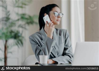 Photo of woman copywriter has telephone talk, looks aside, chats about ideas for new publication, dressed formally, poses in front of laptop computer in coworking space, discuss working process