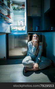 Photo of woman in pajamas eating on floor next to fridge at night