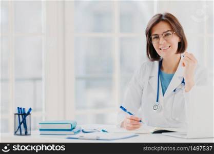 Photo of woman intern works in hospital, writes in medical certificate, prescribes treatment for patient, makes recipe, dressed in white coat with phonendoscope, leaves notes of medical examination