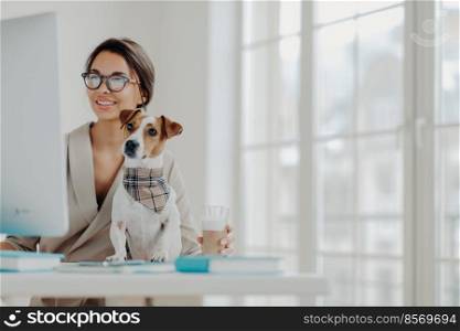 Photo of woman works freelance from home, concentrated in monitor of computer, wears spectacles, poses at desktop with jack russel terrier dog, drinks beverage, smiles positively enjoys favorite work