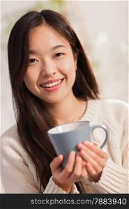 photo of young asian woman with hot mug in her hands smiling towards the camera