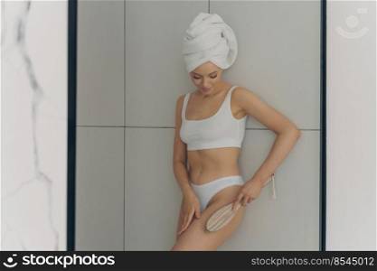 Photo of young beautiful woman doing body massage with dry brush in bathroom after morning shower, wears classic lingerie with head wrapped in towel, isolated on white marble wall background. Photo of young beautiful woman wears classic lingerie doing body massage with brush