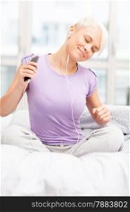 photo of young caucasian woman sitting on a bed and shaking while listening music with headphones