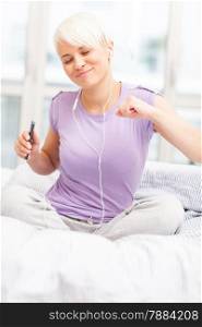 photo of young caucasian woman sitting on a bed and shaking while listening music with headphones