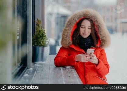Photo of young female in red warm jacket, holds modern cell phone, sends text message to boyfriend, share impessions about winter holidays, stands outdoor during snowing. Technology concept.