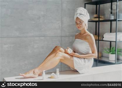Photo of young female model enjoys skin care treatment, applies cream on legs, has healthy smooth glowing skin, well groomed body, poses wrapped in towel in bathroom, uses eco organic cosmetic