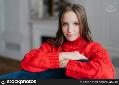 Photo of young good lookng brunette woman enjoys day off at home, has serious look at camera, wears warm loose red sweater, poses at home against blurred background. Coziness and rest concept