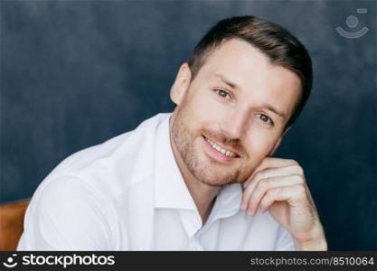 Photo of young male entrepreneur with bristle, keeps hand on cheek, dressed in elegant white shirt, poses against dark background, has toothy smile. Elegant handsome young successful man indoor