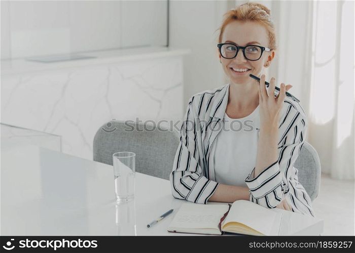 Photo of young red-haired smiling businesswoman using voice assistant app on smartphone, sitting at table at home and planing her day in morning, making notes in notebook and drinking mineral water. Young smiling businesswoman using voice assistant app on smartphone, sitting at table at home