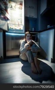 Photo of young woman eating next to refrigerator at night