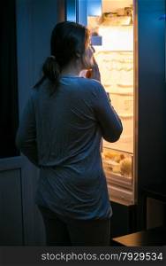 Photo of young woman looking inside the fridge at late night