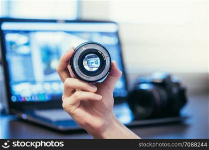 Photographer holds a professional photo optic in his hand. Camera and Laptop in the blurry background.