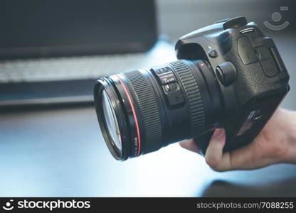 Photographer is holding a professional camera with telephoto lens in his hand, laptop in the blurry background