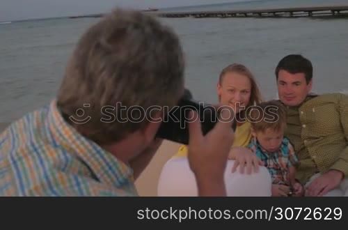 Photographer with professional camera taking shots of happy young family with little son lying on chaise longue at the seaside