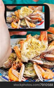 photographing food concept - tourist takes picture of plate with cut crab, oysters, shrimps and other seafood on smartphone, France