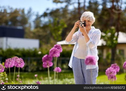 photography, leisure and people concept - happy senior woman with camera photographing flowers blooming at summer garden. senior woman with camera photographing flowers