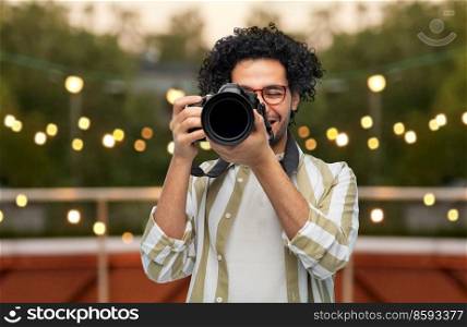 photography, profession and people and concept - happy smiling man or photographer in glasses with digital camera over party lights on roof top background. man or photographer with digital camera at party