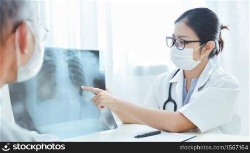 Physician talking and looking at X Ray film with senior man at clinic pandemic virus. Asian woman Doctor wear eyeglasses and surgical mask discussing with Elderly patient in medical room at hospital