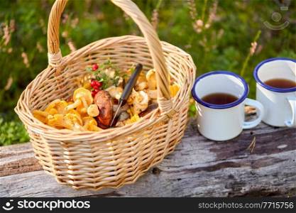picking season and leisure concept - mushrooms in basket and two tin mugs with tea on log in forest. mushrooms in basket and cups of tea in forest