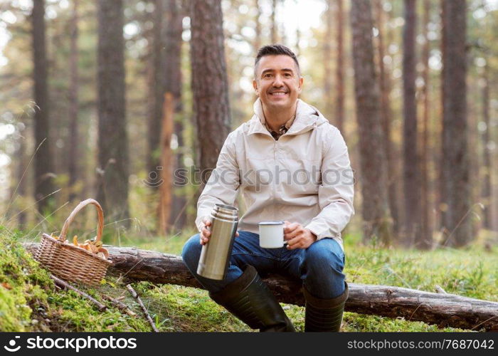 picking season and leisure people concept - happy man with wicker basket of mushrooms sitting on fallen tree and drinking hot tea in autumn forest. man with basket of mushrooms drinks tea in forest