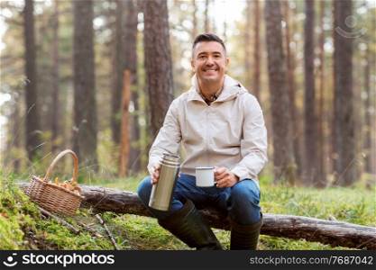 picking season and leisure people concept - happy man with wicker basket of mushrooms sitting on fallen tree and drinking hot tea in autumn forest. man with basket of mushrooms drinks tea in forest