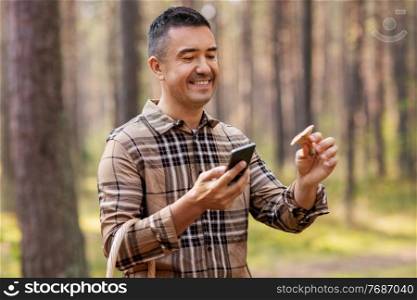 picking season and leisure people concept - happy smiling middle aged man with wicker basket and smartphone using app to identify mushroom in autumn forest. man using smartphone to identify mushroom