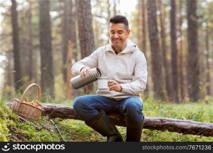 picking season and leisure people concept - smiling man with wicker basket sitting on fallen tree and pouring hot tea to mug in autumn forest. man with basket of mushrooms drinks tea in forest