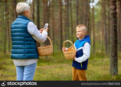 picking season, leisure and people concept - grandmother with smartphone photographing happy smiling grandson with mushrooms in basket in forest. grandmother photographing grandson with mushrooms