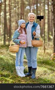 picking season, leisure and people concept - happy smiling grandmother and granddaughter with mushrooms in baskets taking picture with smartphone on selfie stick in forest. grandma with granddaughter taking selfie in forest