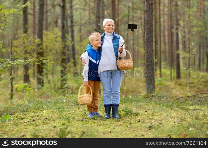 picking season, leisure and people concept - happy smiling grandmother and grandson with mushrooms in baskets taking picture with smartphone on selfie stick in forest. grandmother and grandson with baskets take selfie