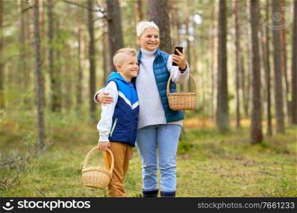 picking season, leisure and people concept - happy smiling grandmother and grandson with mushrooms in baskets taking selfie with smartphone in forest. grandmother and grandson with baskets take selfie