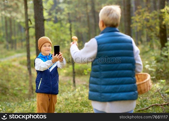 picking season, leisure and people concept - happy smiling grandson with smartphone photographing grandmother with mushroom and basket in forest. grandson photographing grandmother with mushroom