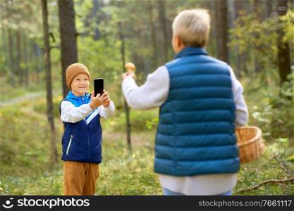 picking season, leisure and people concept - happy smiling grandson with smartphone photographing grandmother with mushroom and basket in forest. grandson photographing grandmother with mushroom