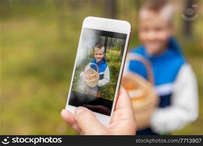 picking season, leisure and people concept - parent with smartphone photographing happy smiling grandson with mushrooms in basket in forest. parent photographing grandson with mushrooms