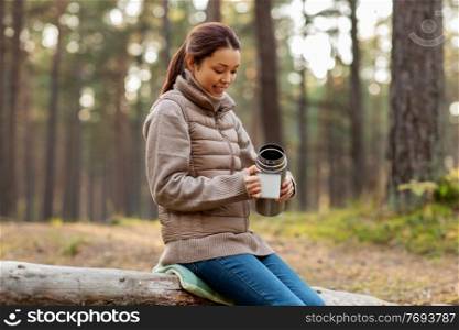picking season, leisure and people concept - young asian woman with thermos and mug drinking tea in autumn forest. asian woman with thermos drinking tea in forest