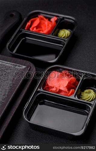 Pickled ginger, soy sauce and wasabi in portioned plastic containers on a dark concrete background