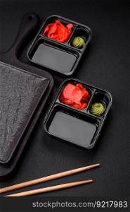 Pickled ginger, soy sauce and wasabi in portioned plastic containers on a dark concrete background