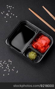 Pickled ginger, soy sauce and wasabi in portioned plastic containers on a dark concrete background