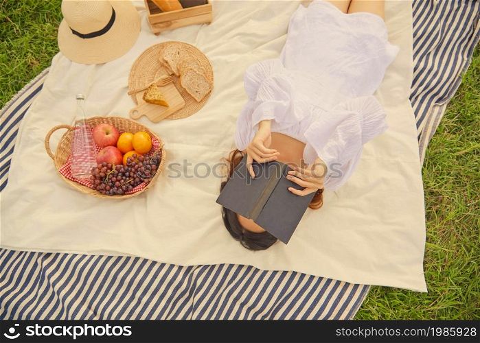 picnic concept The female in the gorgeous white dress sleeping on the white and blue striped cloth with the black book covering face while being on the nice picnic.