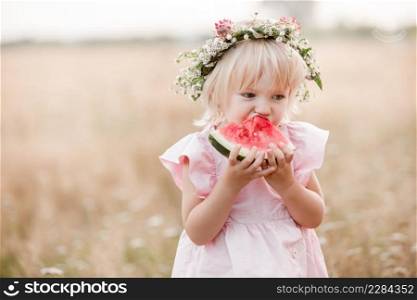 picnic with family. cute little girl eating big piece of watermelon on the grass in summertime in the park. Adorable child wearing in flowers wreath on head and pink dress. picnic with family. cute little girl eating big piece of watermelon on the grass in summertime in the park. Adorable child wearing in flowers wreath on head and pink dress.